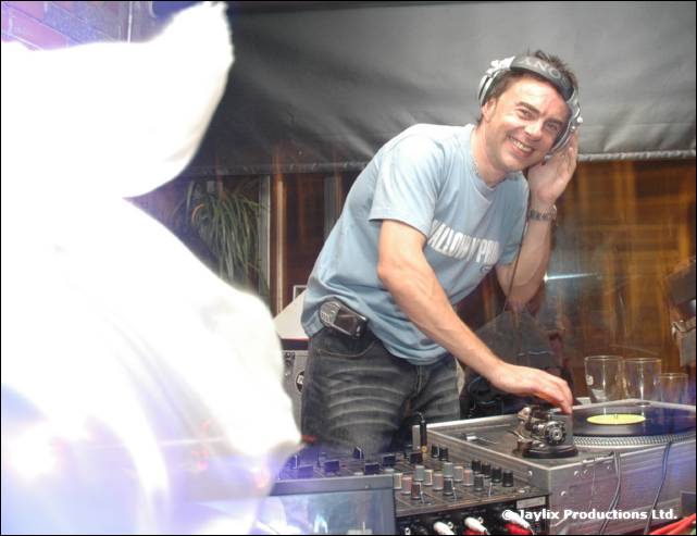 A smiling DJ in headphones and a light blue T-shirt mixes at the decks