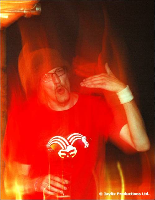 A person in a red T-shirt with a jester logo dances with a raised hand and glasses, in a blur of flames