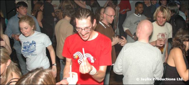 A crowd dances, a man in a red T-shirt with a jester logo laughing and holding a finger up
