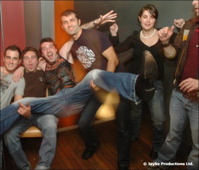 Men at a bar and on a sofa cheer together, one lying across others' laps, in an orange-walled room