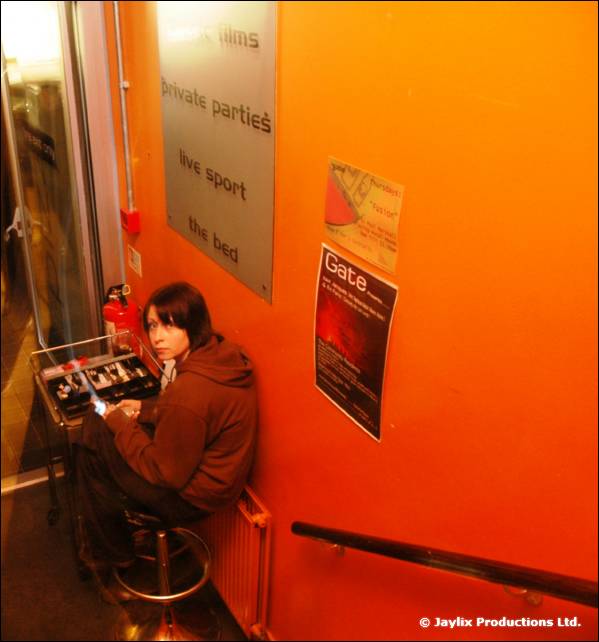 A person sits at a small mixing desk on a stool in an orange-walled stairwell, with a Gate poster