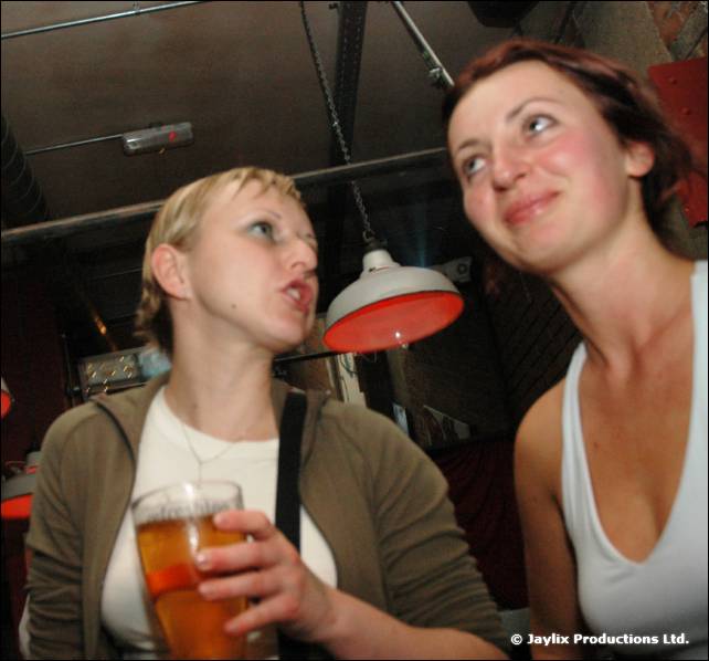 Two women talk and look up, one holding a pint, under a hanging red lamp