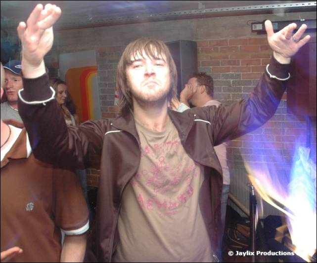 A person with long hair and raised arms dances in a bar, wearing a brown jacket and pink printed T-shirt