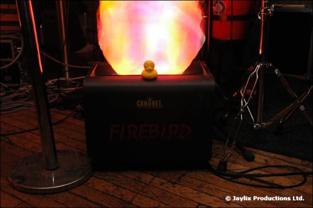 A yellow rubber duck sits on top of a glowing Chauvet Firebird light on a wooden floor
