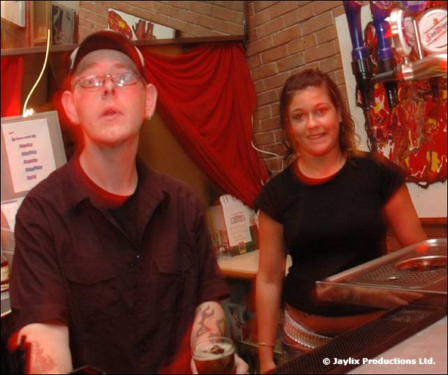 A man in a cap and glasses and a woman in black stand behind a bar with a red curtain and beer taps