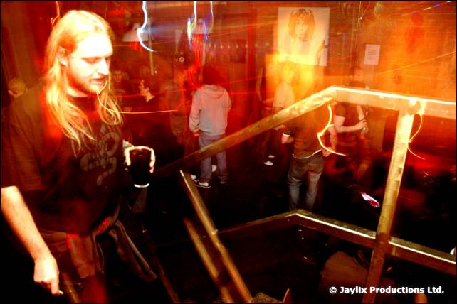 A long-haired man with a beard holds a pint beside a metal stair rail in a dim club