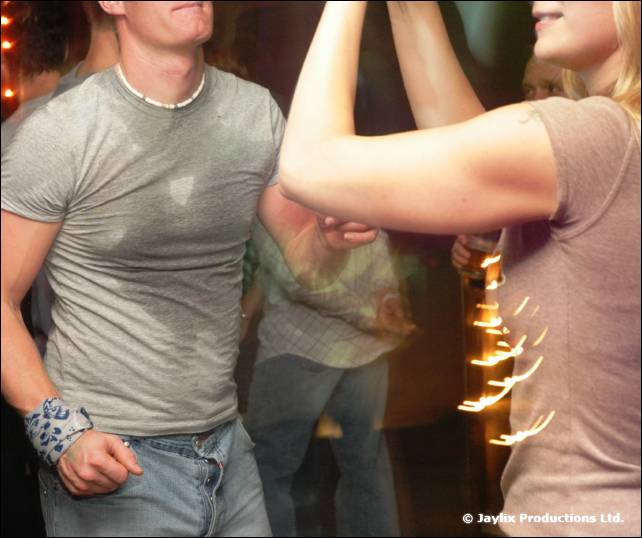 A man in a grey T-shirt dances among other people, a woman's raised arm beside him