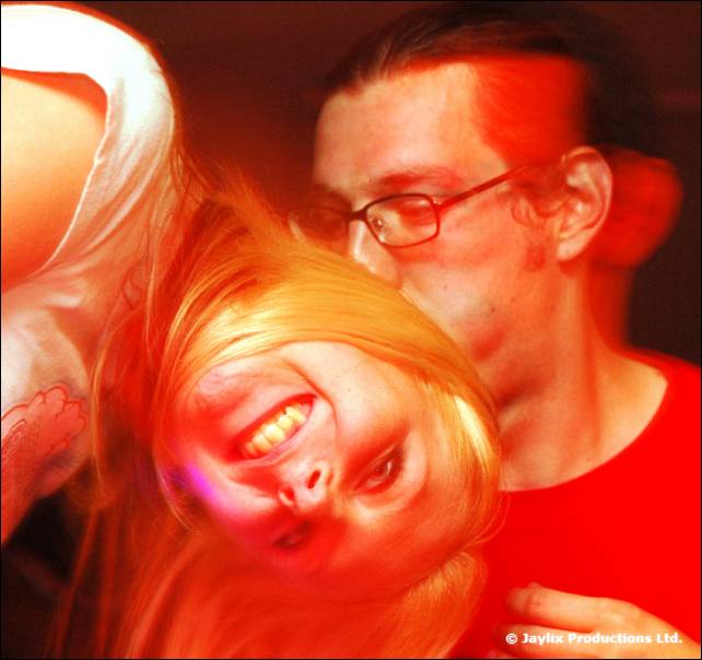 A smiling person with blonde hair tilts their head close to a person with glasses in a red-lit blur