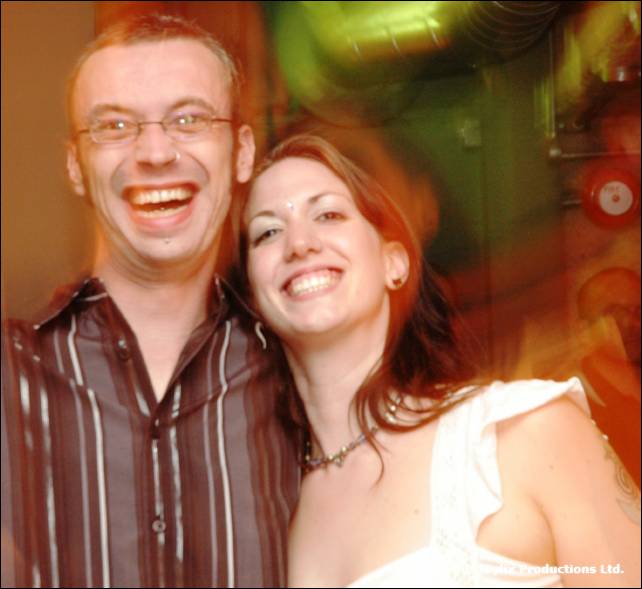 A smiling man with glasses and a woman with dark red hair pose together in a colourful blur