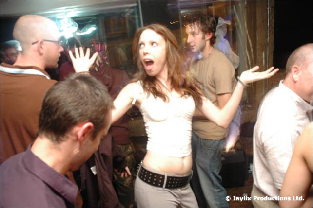 A woman dances with wide-eyed expression and arms out among men in a busy bar