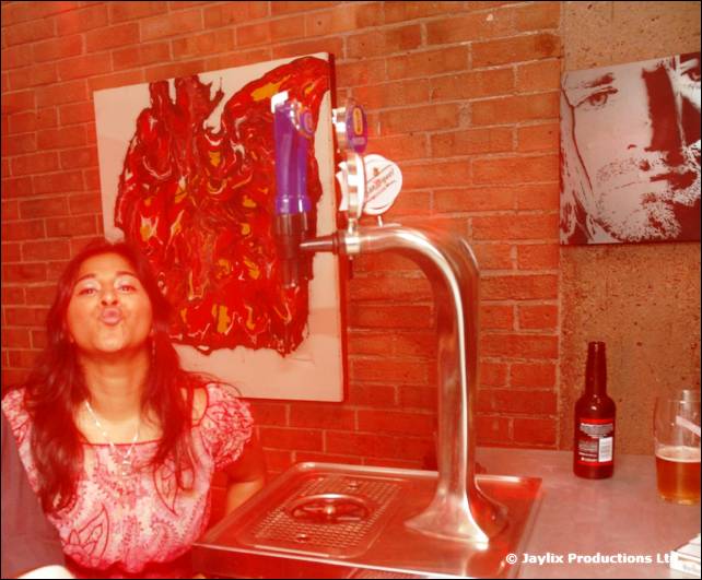 A woman blows a kiss at a bar beside a beer tap, with abstract art and a portrait on the wall