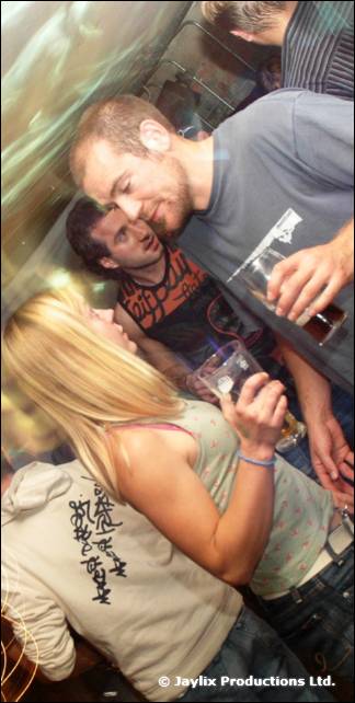 Two men talk closely by a window, one holding a glass, with a woman with blonde hair in the foreground
