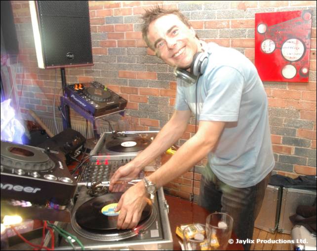 A smiling DJ in a light blue T-shirt and headphones mixes at Pioneer decks and turntables