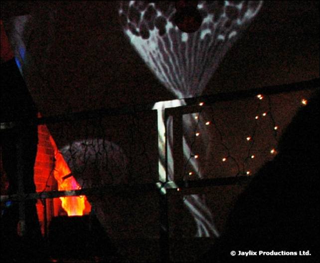 A dark projection on a wall with a small orange flame and string lights on a railing