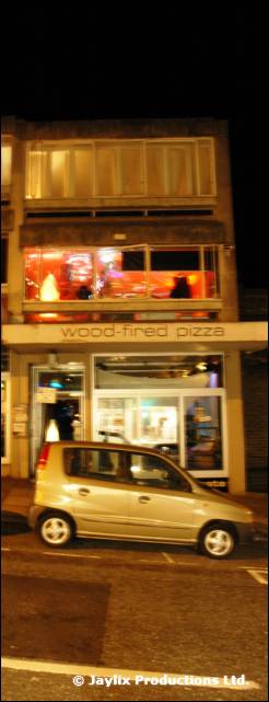 A pizza restaurant front at night with a silver car parked outside, sign reading wood-fired pizza