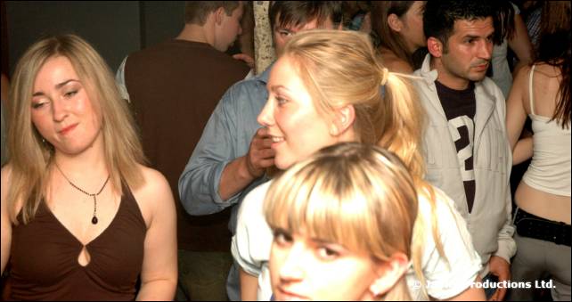 A woman with blonde hair and several people dance and talk in a crowded bar