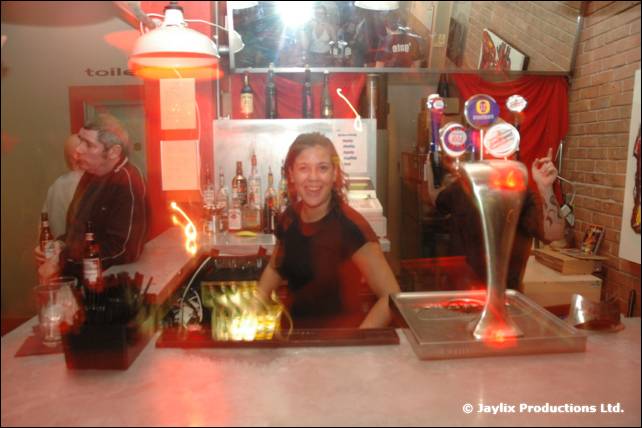 A woman serves behind a bar with a tall beer tap and bottles, lit warmly