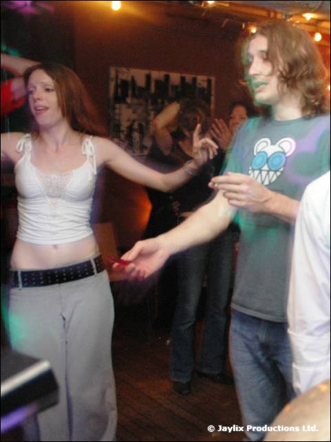 A woman in a white top and a man in a band T-shirt dance together in a bar