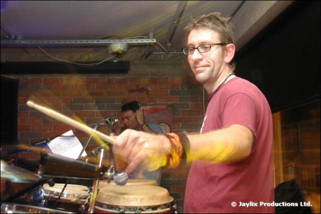 A man with glasses holds drumsticks over a conga drum in a bar