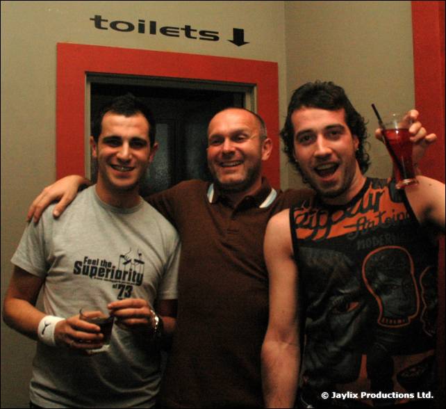 Three men pose with arms round each other and drinks beside a sign reading toilets