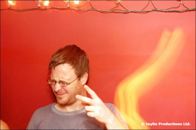 A man with glasses pulls a face and holds up two fingers against a red wall