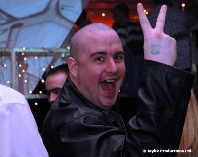 A bald person in a black leather jacket grins and holds up a peace sign