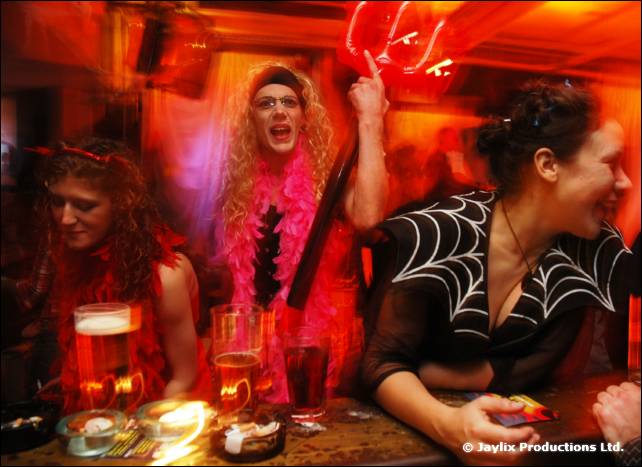 A person in a pink feather boa and others sit at a bar table with pints, one woman in a spider web top