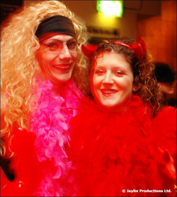 Two people in fancy dress, one in a blonde wig and pink boa, one in devil horns and a red feather boa