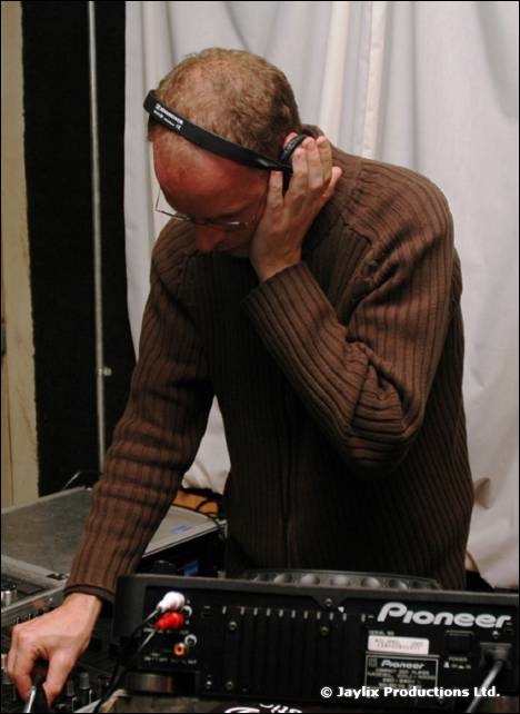 A DJ in a brown jumper and headphones leans over Pioneer equipment in front of white drapes