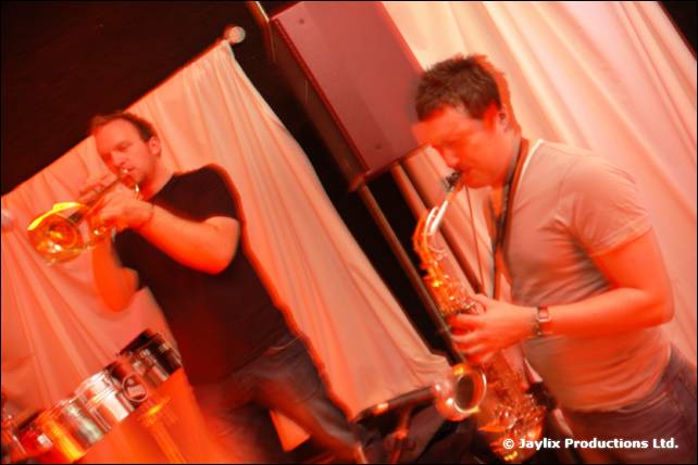 Two musicians play a trumpet and a saxophone in front of white drapes