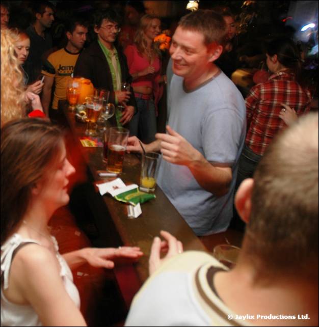 People talk and drink around a bar table in a busy pub, one woman in a white top