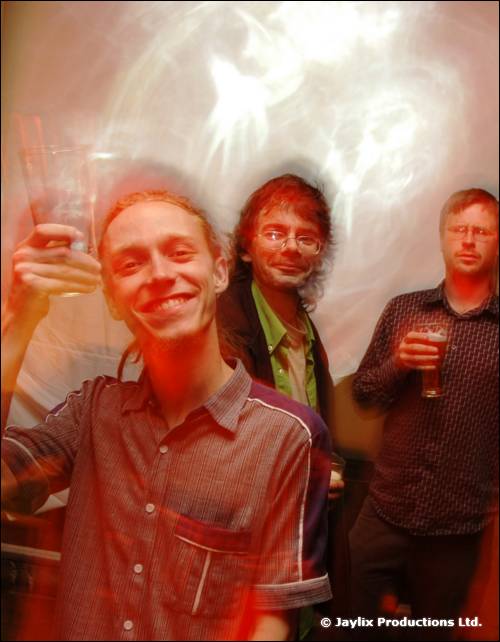 Three men stand together smiling, one raising a glass, against a hazy white background