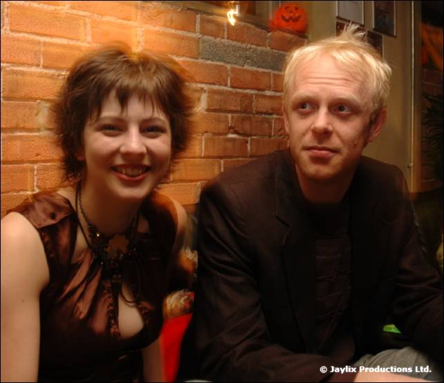 A woman and a man with blonde hair sit close together by a brick wall with a pumpkin