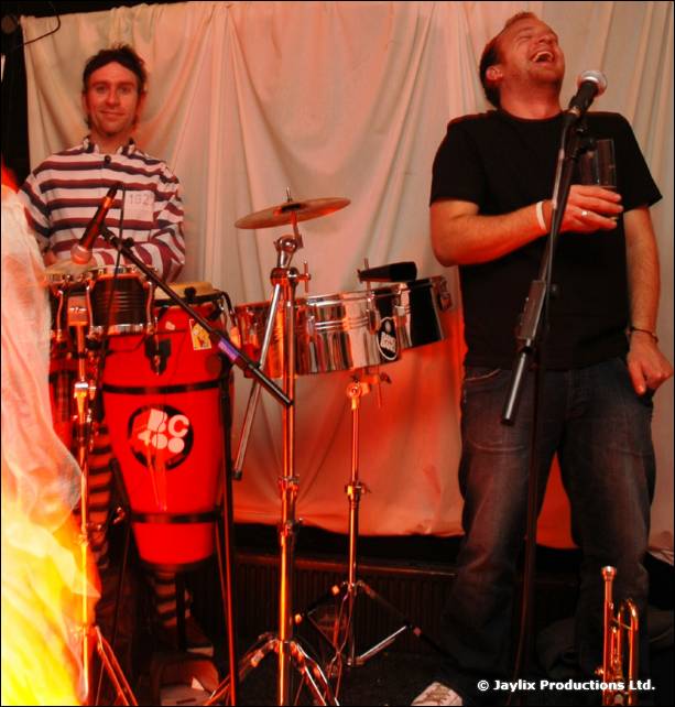A drummer and a singer at a microphone perform in front of white drapes, with congas and a drum kit
