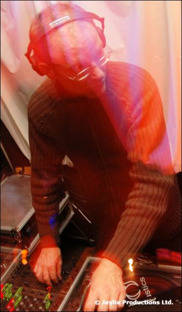 A DJ in headphones and brown jumper mixes at a mixing desk, with motion blur