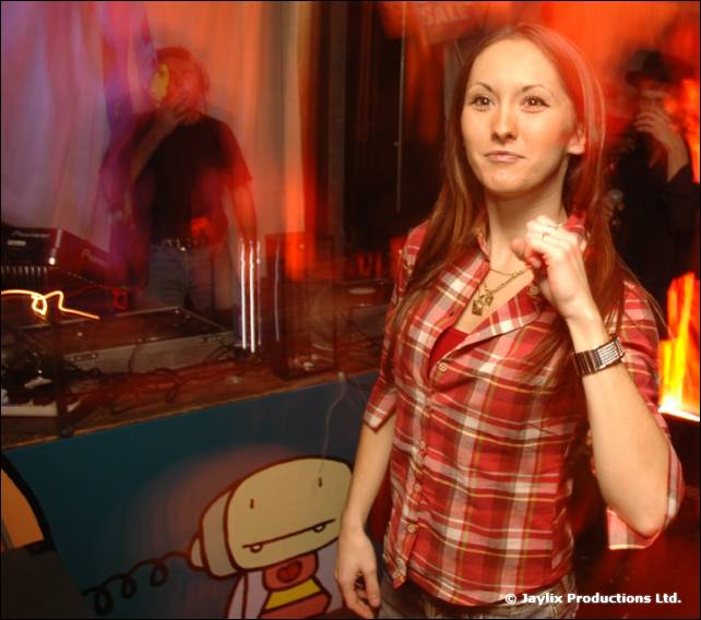 A woman in a red check shirt stands in front of DJ decks with a blurred DJ behind her