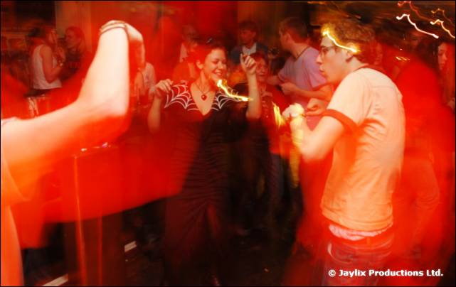 A woman in a black dress with a spider-web pattern dances in a red-lit crowd, a man in an orange t-shirt beside her