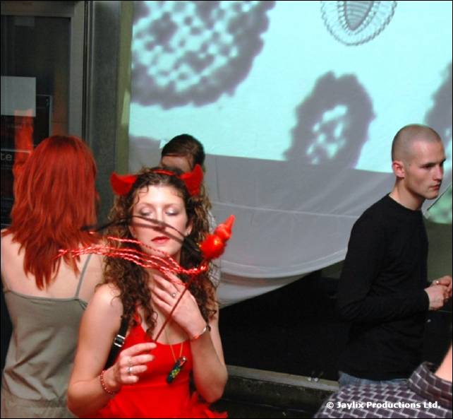 A woman in a red dress with devil horns holds a toy trident, with a red-haired woman and a man beside her