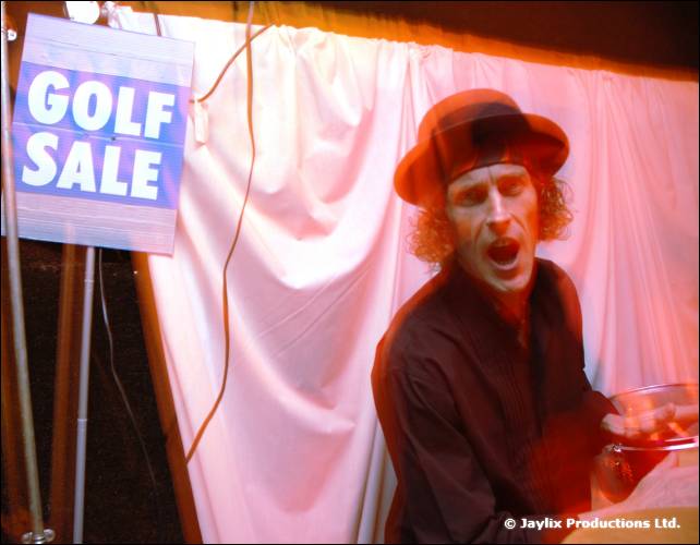 A man in a brimmed hat holds a drink and shouts beside a white curtain and a Golf Sale sign