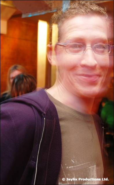 A smiling man in glasses and a purple hoodie leans towards the camera in a dim bar