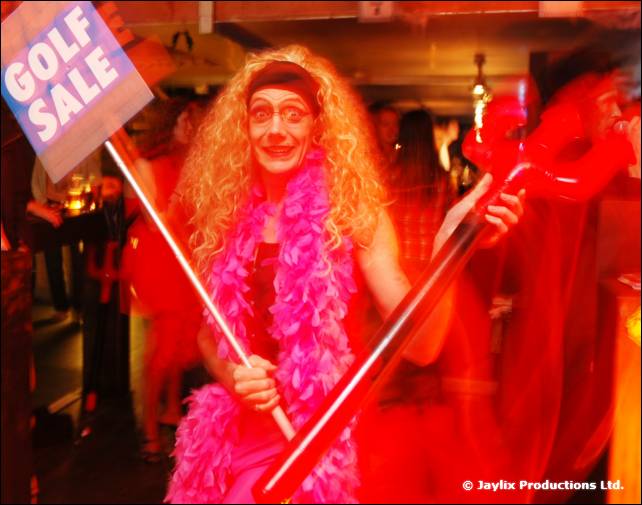 A person in a long curly blonde wig and pink feather boa holds a Golf Sale sign and a red toy trident
