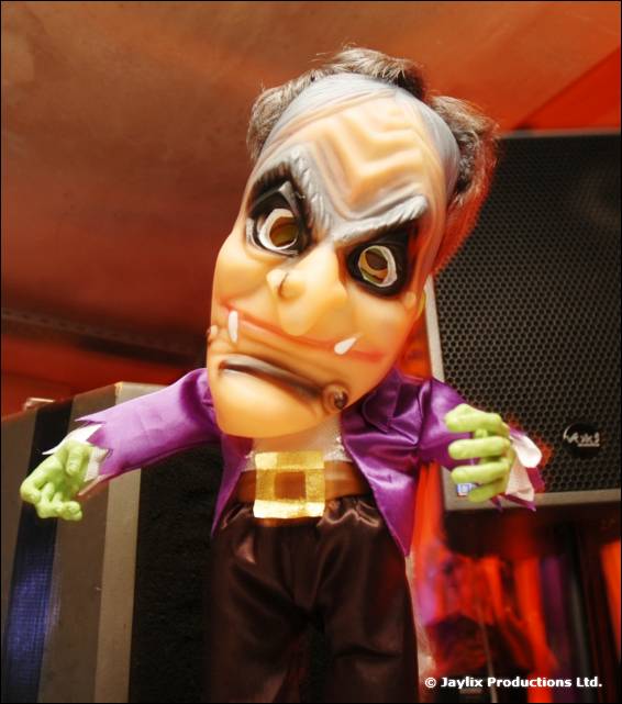 A large grotesque mask puppet with a white wig, purple jacket and green hands stands on a speaker
