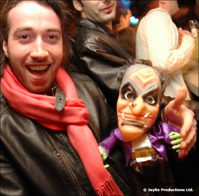 A smiling man in a leather jacket and red scarf holds up a grotesque puppet in a crowd