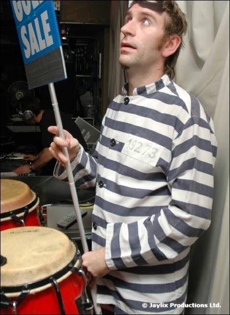 A man in a striped prisoner costume holds a Golf Sale sign beside conga drums