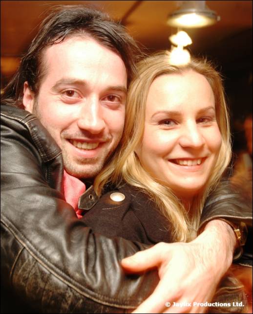 A man in a leather jacket hugs a blonde woman from behind as they smile