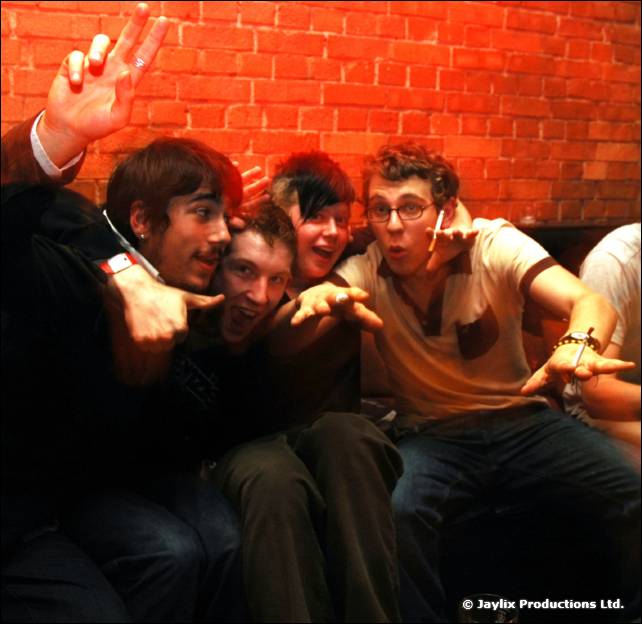 Five people crowd together against a red brick wall, pulling faces and making hand signs