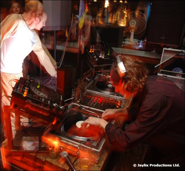 A DJ in headphones uses Pioneer decks while another person stands behind in a white top