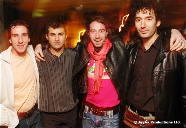 Four men stand together with arms around each other, one in a pink scarf and leather jacket