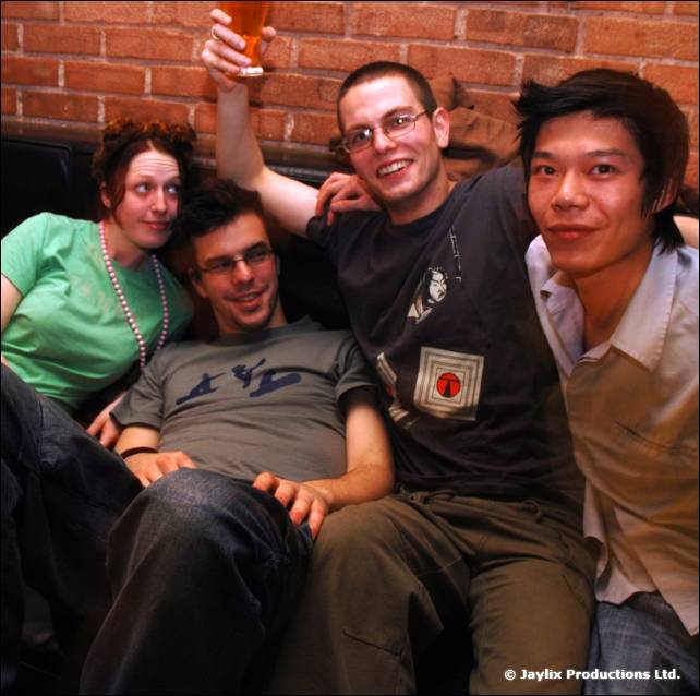 Five people sit squeezed together on a sofa against a brick wall, one raising a beer