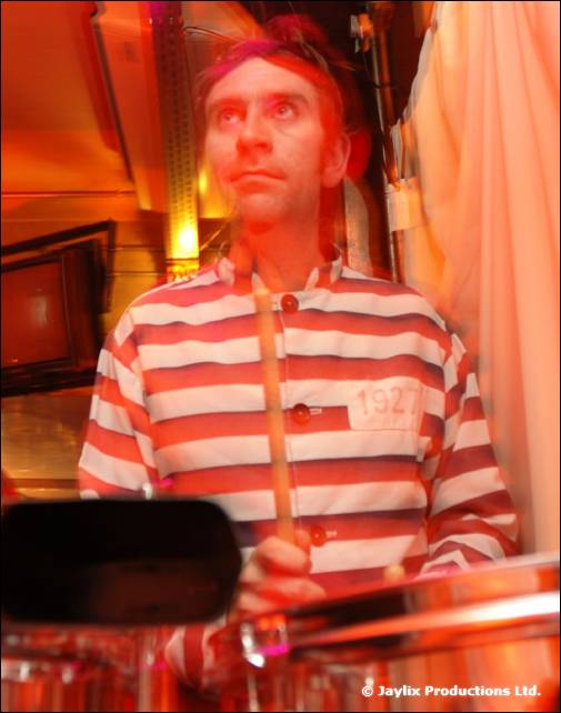 A person in a red and white striped prisoner top with a headband holds a drumstick over a drum